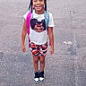 child, girl, smiling, outdoor, pavement, braided_hair, beads, tshirt, cartoon_print, shorts, camouflage, sneakers, backpack, standing, happy, person, casual_clothing, daylight, street, fashion