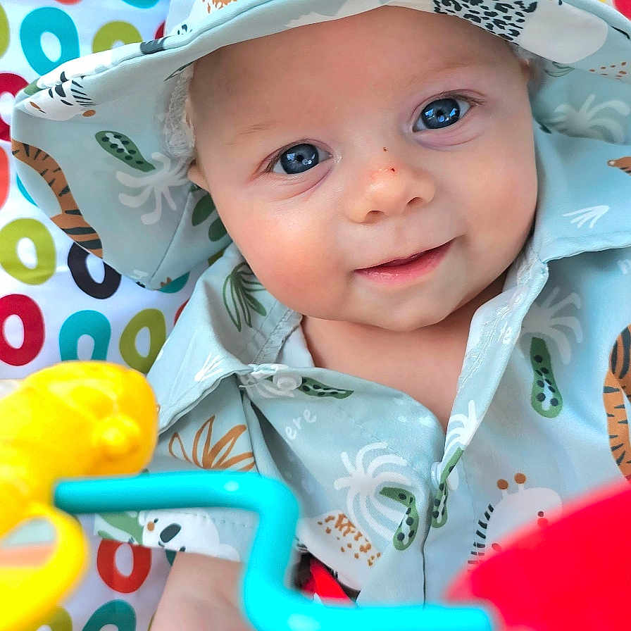 William-Harvey is registered to the contest to win money with this photo: animal_print, baby, blue_eyes, bright, child, closeup, clothing, colorful, cute, face, happy, hat, indoors, infant, patterned_background, playful, portrait, shirt, smile, toy