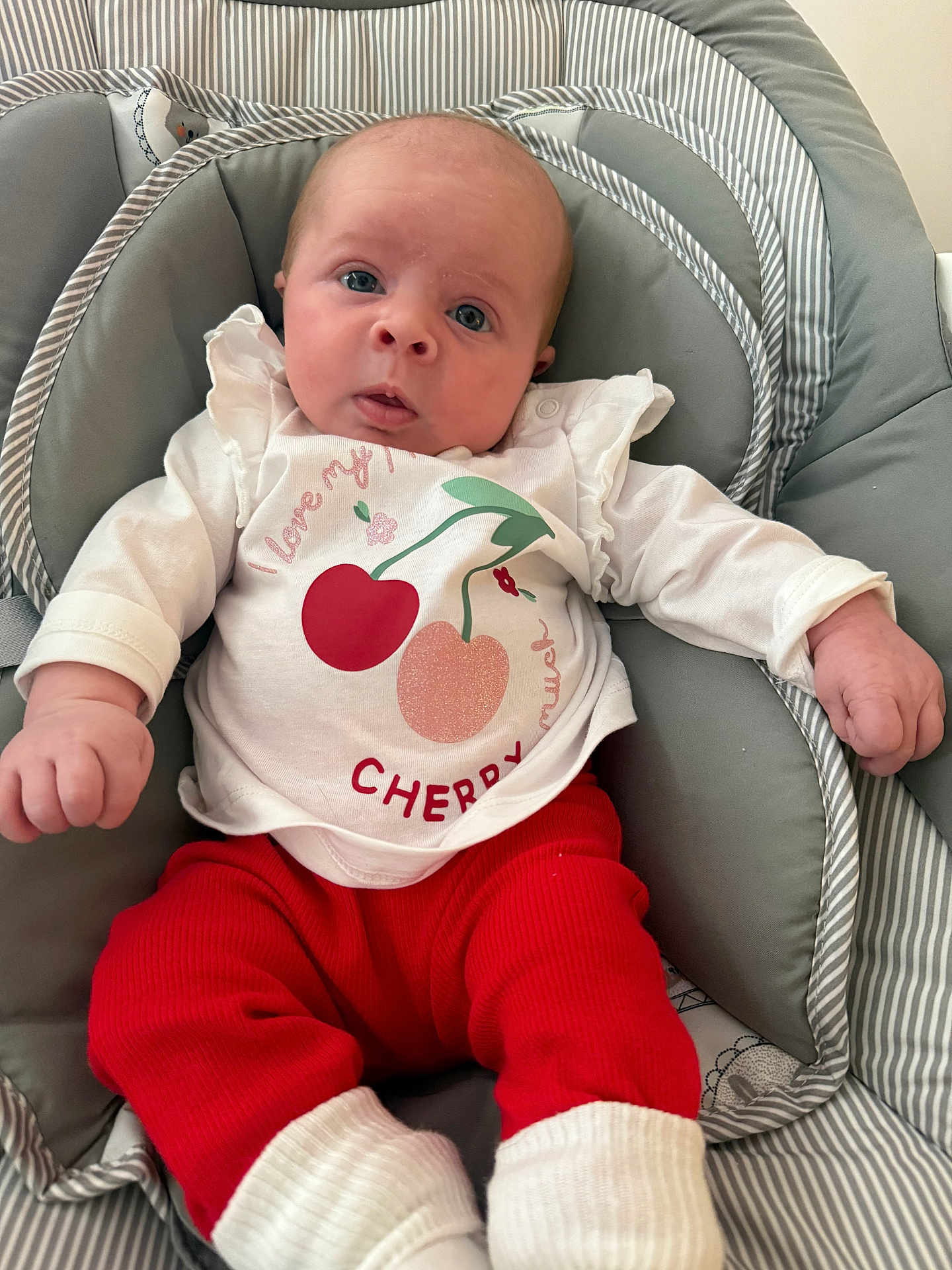 Sophia is registered to the contest to win money with this photo: baby, infant, child, cute, clothing, red_pants, white_shirt, cherry_graphic, cushion, seat, indoor, portrait, small_hands, wide_eyes, expression, cozy, striped_pattern, soft_fabric, sitting, young