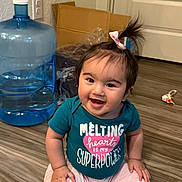 Isabella is registered to the contest to win money with this photo: baby, cardboard_box, child, clothing, cute, door, face, floor, hair_bow, hand, happy, indoor, person, pink_pants, plastic_wrap, sitting, smiling, teal_shirt, water_jug, wooden_floor