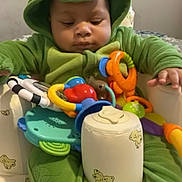 Malik joined the competition — help win amazing prizes! baby, child, dinosaur_onesie, green_clothing, toy, colorful_toys, baby_chair, teddy_bear_prints, indoor, infant, cute, focused, sitting, plush, hand, face, hood, onesie, playtime, adorable