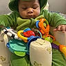 Malik is registered to the contest to win money with this photo: baby, child, dinosaur_onesie, green_clothing, toy, activity_center, baby_seat, teddy_bear_prints, indoor, cute, focused, infant, plush, colorful_toys, hands, sitting, young_child, hooded_clothing, playtime, soft_fabric
