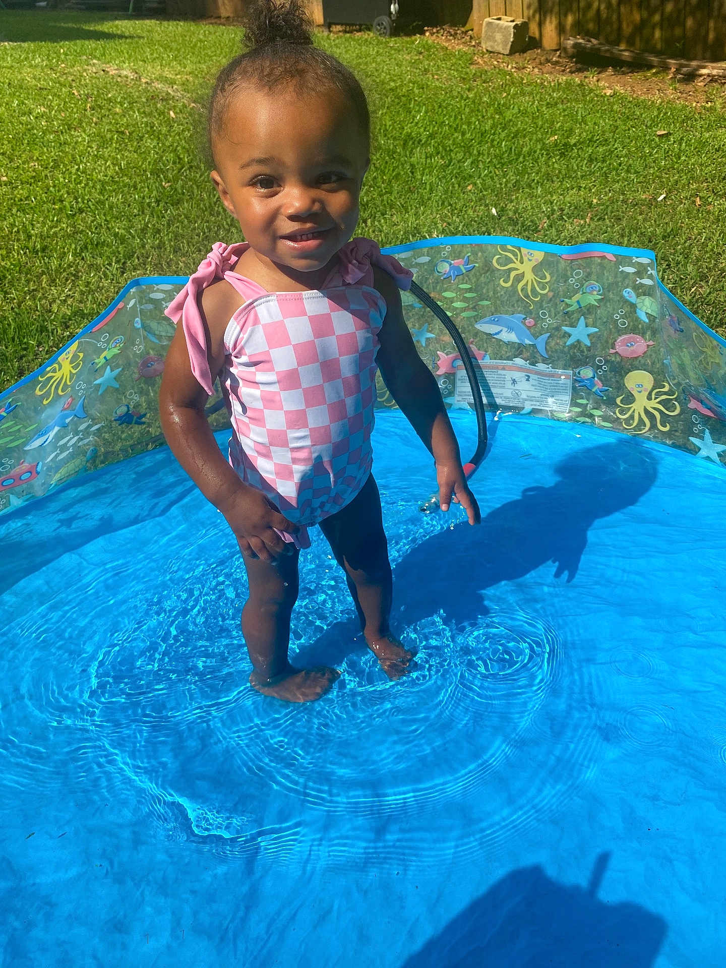 Zuri joined the competition — help win amazing prizes! toddler, child, pool, water, swimsuit, pink, checkered, summer, outdoor, grass, sunlight, happy, smiling, barefoot, splashing, play, backyard, daytime, young_child, person