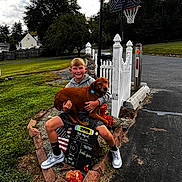Wishbone joined the competition — help win amazing prizes! boy, dog, outdoor, grass, basketball_hoop, white_fence, brick_border, sidewalk, sign, smiling, casual_clothing, tree, cloudy_sky, shoes, shorts, holding_pet, pet, child, happy, summer