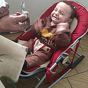Livio a rejoint le concours — aidez-le/la à gagner de superbes lots ! baby, baby_clothes, child, clothing, cute, expression, feeding, floor_tiles, furniture, hand, happy, indoor, infant, joy, person, playful, rocker_chair, seat, smiling, spoon