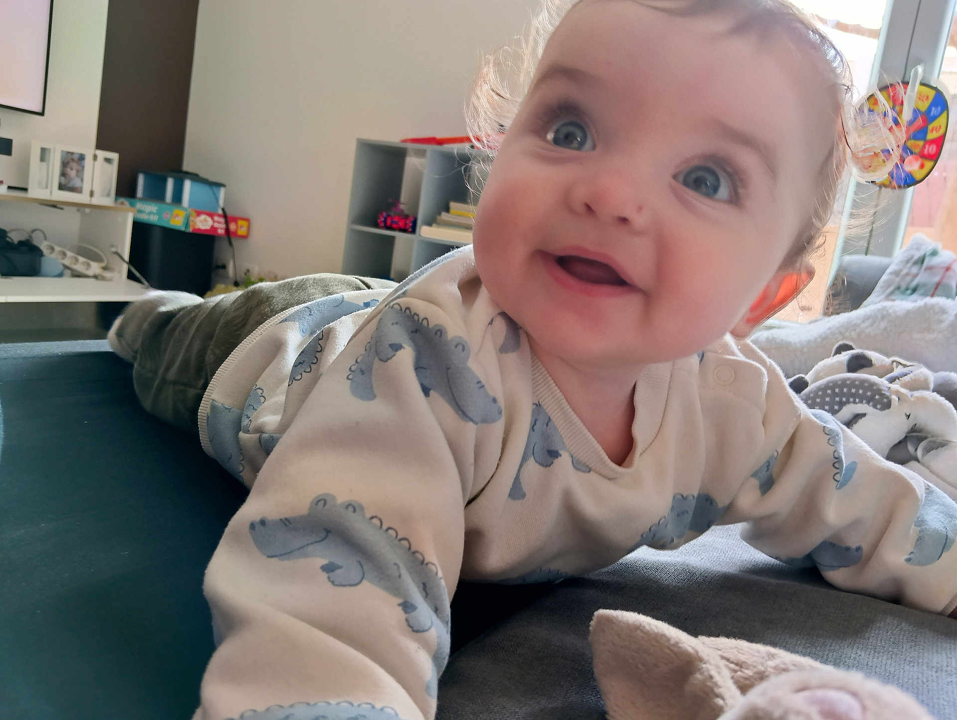 Théo participe au concours pour gagner de l'argent avec cette photo : baby, child, smiling, blue_eyes, curly_hair, sweater, alligator_print, plush_toy, indoor, couch, blanket, happy, portrait, closeup, soft_lighting, home, furniture, floor, person, toddler