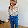 child, girl, braids, pink_bow, glasses, floral_top, denim_pants, sneakers, indoor, wooden_floor, white_wall, standing, pose, casual_clothing, young, portrait, fashion, footwear, cute, expression
