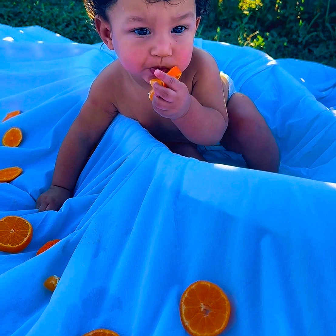 Kadel is registered to the contest to win money with this photo: baby, blue, child, citrus, curly_hair, cute, eating, fence, fruit, garden, inflatable_pool, orange, outdoor, pool, portrait, sitting, snack, summer, sunlight, toddler