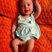 Logan is registered to the contest to win money with this photo: baby, infant, smiling, happy, child, portrait, clothing, dress, diaper, orange_blanket, texture, skin, face, arm, legs, lying_down, cute, indoors, person, young_child