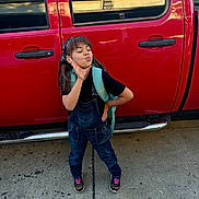 Isabella is registered to the contest to win money with this photo: child, girl, posing, denim_overalls, backpack, black_shirt, red_truck, sidewalk, playful, outdoor, casual_clothing, sneakers, headband, pigtails, concrete, vehicle, reflection, window, door_handle, expression