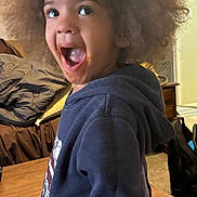 Julius joined the competition — help win amazing prizes! child, afro_hair, hoodie, indoor, expression, messy_face, table, furniture, carpet, person, surprise, excitement, young, casual_clothing, living_room, smile, mouth_open, hair, curly_hair, home