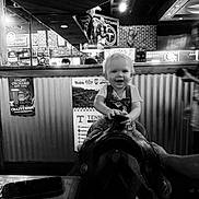 Whyatt joined the competition — help win amazing prizes! baby, child, toddler, smiling, bow_tie, mechanical_bull, ride, indoor, sports_bar, restaurant, black_and_white, portrait, seated, happy, blond_hair, face, furniture, brick_wall, signs, entertainment