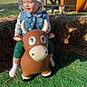 Whyatt is registered to the contest to win money with this photo: child, toddler, baby, riding_toy, inflatable_cow, cow, animal_toy, cat_hat, sun_hat, denim_vest, green_shirt, socks, outdoor, farm, hay_bales, picnic_table, kayaks, countryside, sunlight, portrait