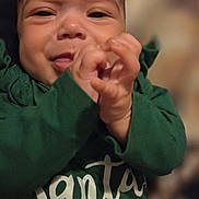 Amelia joined the competition — help win amazing prizes! baby, child, smile, face, hand, green_clothing, sweater, text_on_clothing, portrait, indoors, cute, infant, person, closeup, happy, skin, young_child, warm_lighting, background_blur, holding