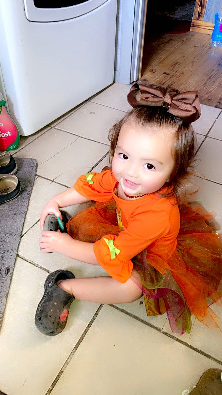 Delaney is registered to the contest to win money with this photo: baby_toddler_clothing, child, floor, flooring, foot, fun, hairstyle, happy, human_leg, mobile_phone, person, pink, room, sandal, sitting, smile, sock, thigh, thumb, toddler