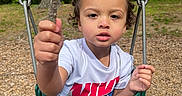 Kawhi is registered to the contest to win money with this photo: toddler, child, swing, playground, stick, outdoor, trees, woodchips, green_swing, curly_hair, white_shirt, blue_pants, nature, daylight, person, holding, expression, young_child, park, casual_clothing