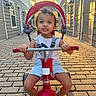 Kawhi is registered to the contest to win money with this photo: toddler, child, tricycle, outdoor, sunlight, brick_path, curly_hair, smiling, white_shirt, shorts, black_shoes, residential_area, door, window, daytime, play, toy, person, happy, young_child