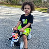 child, toddler, tricycle, outdoor, street, toy, mr_potato_head, curly_hair, black_shirt, yellow_shorts, sneakers, daylight, trees, grass, asphalt, play, recreation, kid, person, summer