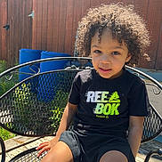 Kentrell is registered to the contest to win money with this photo: child, curly_hair, black_shirt, reebok, shorts, metal_chair, outdoor, sunlight, wooden_fence, blue_bins, summer, smiling, person, portrait, daylight, casual, backyard, seat, happy, young