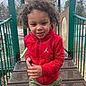 child, curly_hair, red_hoodie, playground, outdoor, smiling, person, young, happy, metal_stairs, green_rails, trees, overcast_sky, casual_clothing, daytime, park, active, cute, motion, play