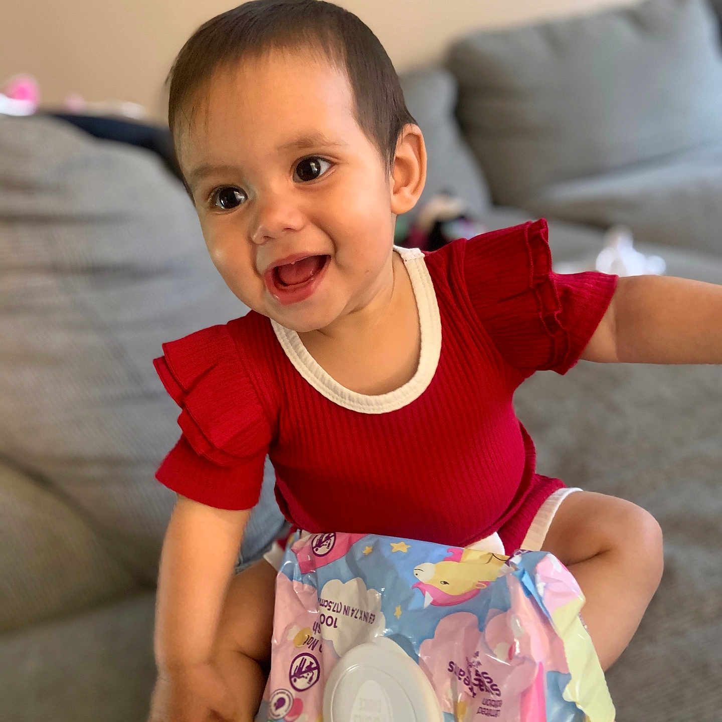 Legacie joined the competition — help win amazing prizes! baby, child, smiling, red_clothing, sitting, couch, indoor, happy, face, infant, skin, arm, hand, package, baby_wipes, colorful, toy, young_child, portrait, casual