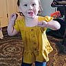 Malaya joined the competition — help win amazing prizes! toddler, child, yellow_top, blue_pants, barefoot, carpet, indoor, playful, tongue_out, person, young_child, hair_tied, furniture, door, household, living_room, rug, standing, cute, smiling