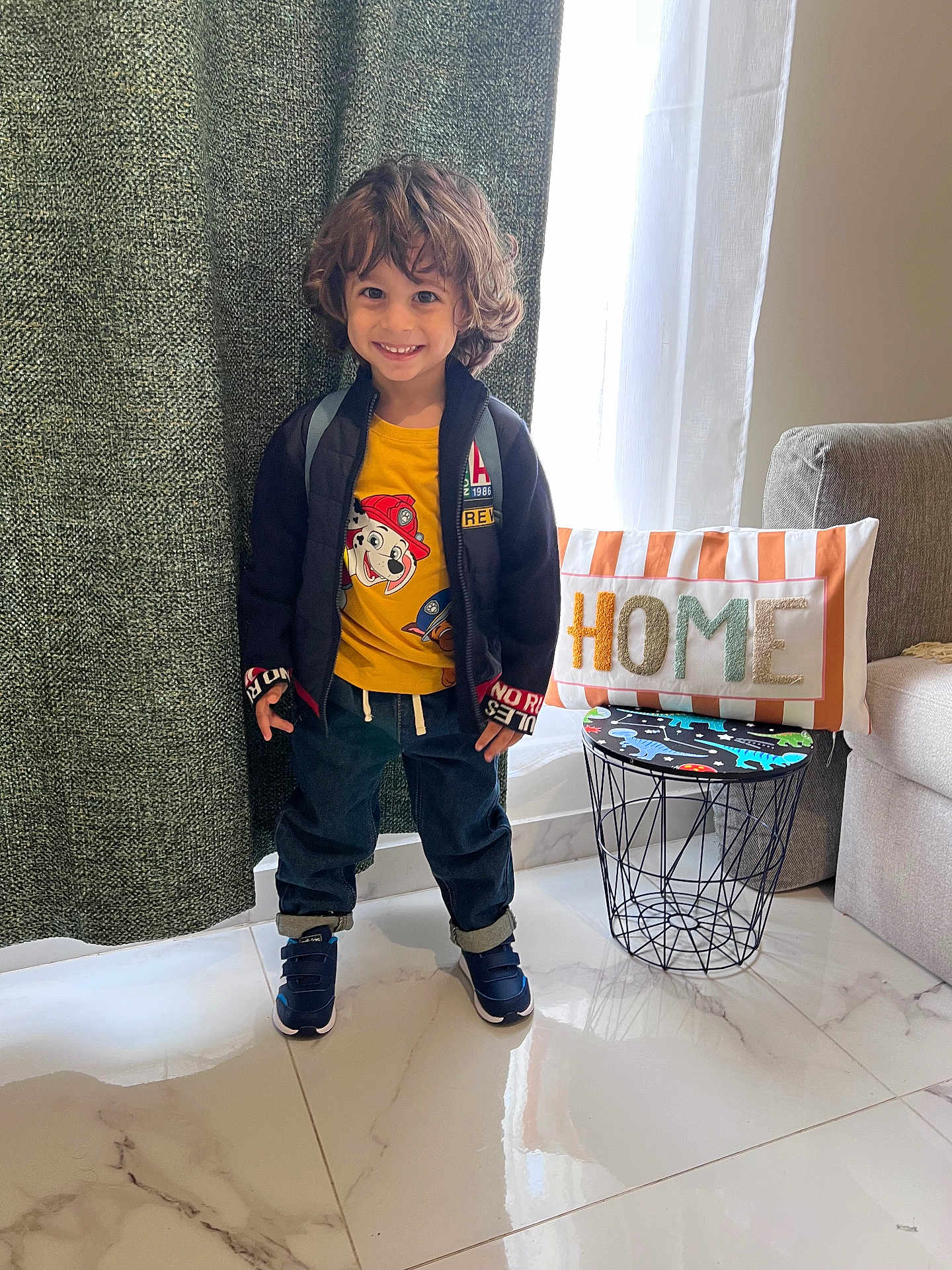 Moussa participe au concours pour gagner de l'argent avec cette photo : child, smiling, indoor, curly_hair, yellow_tshirt, jacket, jeans, sneakers, backpack, pillow, home_decor, table, window, curtain, floor, marble_floor, living_room, person, cute, happy