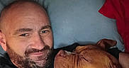 Mona is registered to the contest to win money with this photo: man, dog, bed, pillow, blanket, indoor, portrait, smile, beard, shaved_head, brown_dog, snuggling, relaxed, closeup, friendship, cozy, casual, resting, human, pet