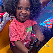 Xianah joined the competition — help win amazing prizes! child, smiling, curly_hair, arcade, steering_wheel, pink_shirt, indoor, play_area, excitement, seat, yellow, black_steering_wheel, shorts, person, fun, game, colorful, happy, amusement, entertainment