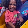 child, smiling, curly_hair, arcade, steering_wheel, pink_shirt, indoor, play_area, excitement, seat, yellow, black_steering_wheel, shorts, person, fun, game, colorful, happy, amusement, entertainment