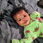 Josiah joined the competition — help win amazing prizes! baby, child, infant, smiling, green_clothing, football_pattern, onesie, curly_hair, lying_down, blanket, gray_blanket, happy, cute, indoor, portrait, person, young_child, face, expressive_eyes, cozy