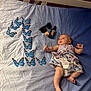 baby, infant, bed, butterfly, blue, dress, sunglasses, flower, bottle, pattern, lying_down, cute, skin, hand, foot, fabric, indoor, decor, arrangement, peaceful