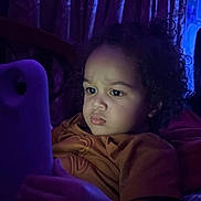 Soren joined the competition — help win amazing prizes! child, toddler, curly_hair, face, portrait, sitting, indoors, tablet, screen, technology, purple_lighting, blue_lighting, curious, focused, serious_expression, clothing, shirt, curtains, couch, home