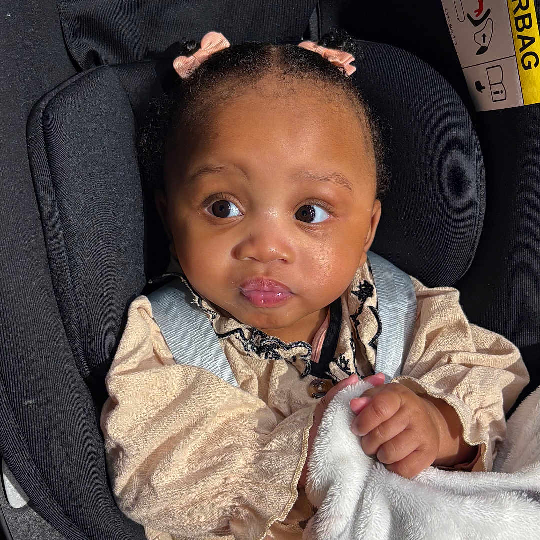 Khadidja Ifeoma a rejoint le concours — aidez-le/la à gagner de superbes lots ! adorable, baby, blanket, car_seat, child, clothing, curly_hair, cute, expression, indoor, infant, light_and_shadow, looking_away, pink_bows, portrait, safety, seatbelt, sitting, soft_texture, transportation