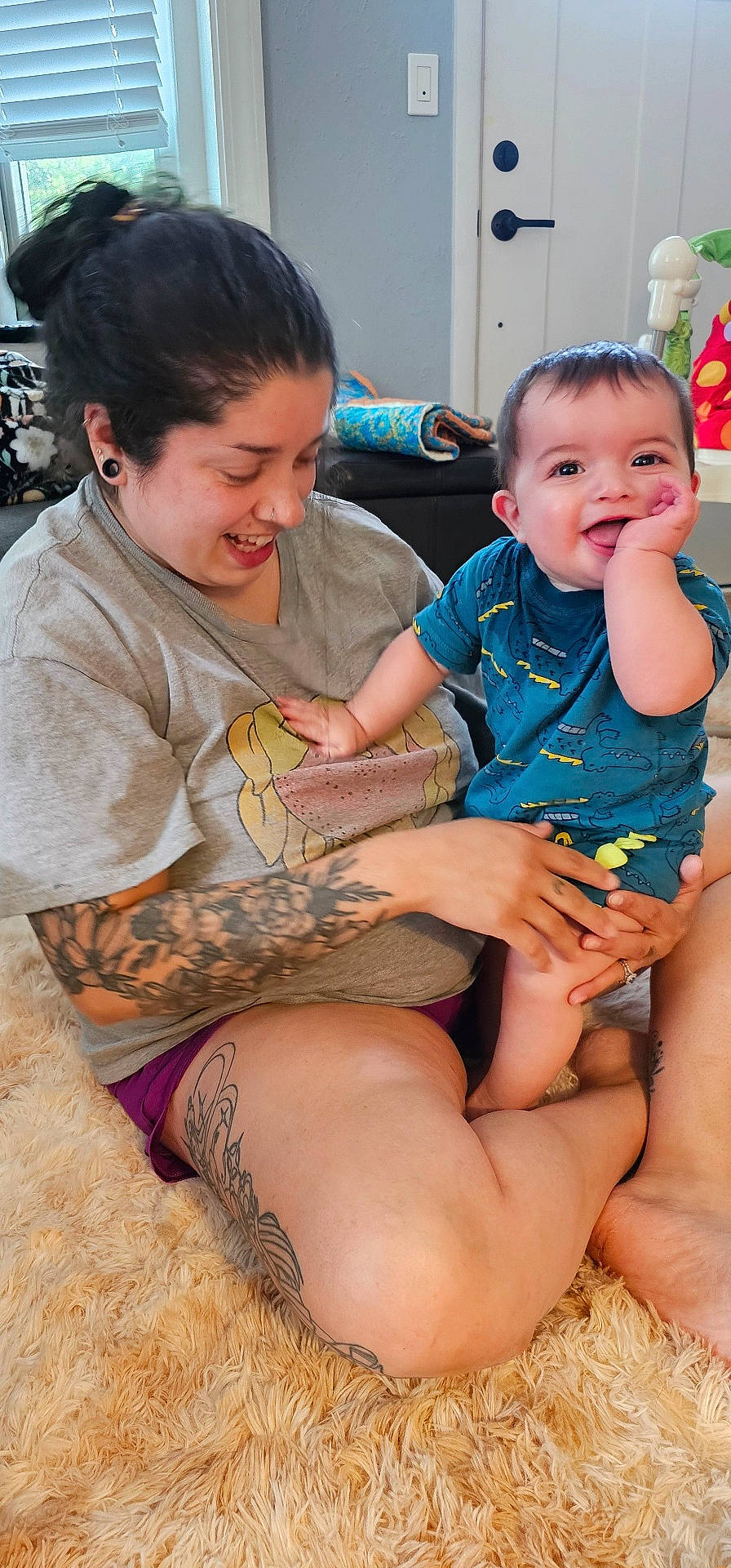 Beck joined the competition — help win amazing prizes! baby, blinds, carpet, child, door, furniture, hand, happy, indoor, living_room, person, playful, rug, shorts, sitting, smiling, tattoos, tshirt, window, woman