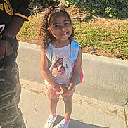 Bradlee joined the competition — help win amazing prizes! child, girl, smiling, curly_hair, backpack, sunlight, sidewalk, tree, bushes, casual_clothing, shorts, sneakers, pink_socks, person, outdoor, nature, happy, portrait, standing, adult_partial