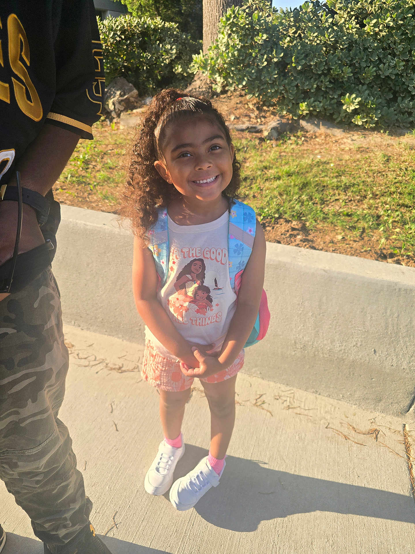 Bradlee joined the competition — help win amazing prizes! child, girl, smiling, curly_hair, backpack, sunlight, sidewalk, tree, bushes, casual_clothing, shorts, sneakers, pink_socks, person, outdoor, nature, happy, portrait, standing, adult_partial