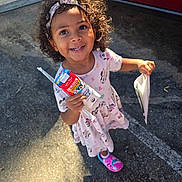 Maddox is registered to the contest to win money with this photo: child, girl, curly_hair, headband, milk_carton, dress, minnie_mouse, pink, shoes, outdoor, sunlight, pavement, car, red_car, smile, happy, person, holding, paper_bag, cute
