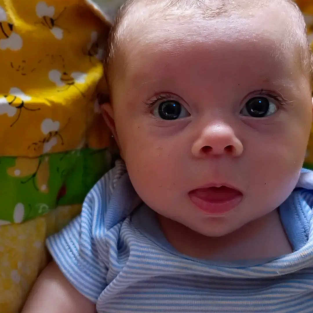 Selah is registered to the contest to win money with this photo: baby, infant, child, blue_onesie, striped_clothing, face, eyes, curious_expression, quilt, colorful_blanket, bee_pattern, arm, skin, portrait, lying_down, indoor, cute, young, person, head