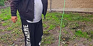 Chica is registered to the contest to win money with this photo: man, puppy, dog, leash, grass, leaves, building, jacket, pants, sneakers, outdoor, smiling, pet, harness, casual_clothing, happy, walking, daylight, nature, sidewalk
