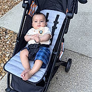 Yuvraj joined the competition — help win amazing prizes! baby, stroller, child, outdoor, sidewalk, brick_wall, concrete, gravel, blue_shorts, white_shirt, infant, person, footwear_free, seat_belt, black_stroller, curious_expression, relaxing, summer_clothing, casual, daytime