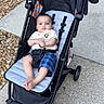 baby, stroller, child, outdoor, sidewalk, brick_wall, concrete, gravel, blue_shorts, white_shirt, infant, person, footwear_free, seat_belt, black_stroller, curious_expression, relaxing, summer_clothing, casual, daytime
