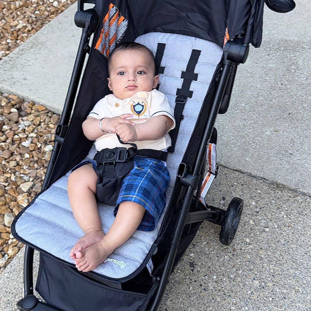 Yuvraj joined the competition — help win amazing prizes! baby, stroller, child, outdoor, sidewalk, brick_wall, concrete, gravel, blue_shorts, white_shirt, infant, person, footwear_free, seat_belt, black_stroller, curious_expression, relaxing, summer_clothing, casual, daytime