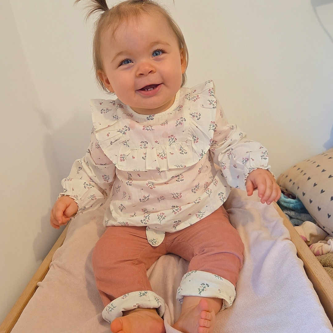 Mya Ladune participe au concours pour gagner de l'argent avec cette photo : baby, barefoot, blanket, changing_table, child, cute, floral_clothing, furniture, happy, indoor, infant, person, pigtails, pink_pants, portrait, room, sitting, smiling, soft_lighting, wooden_frame