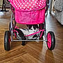 cat, stroller, pink, polka_dot, mesh_basket, wooden_floor, indoor, pet, animal, black_and_white, curious, relaxed, wheel, fabric, furniture, home, cozy, cute, resting, floor