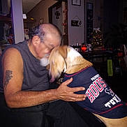 Schatzi is registered to the contest to win money with this photo: man, dog, indoor, affection, blur, tattoo, sleeveless_shirt, houston_texans, pet_clothing, embrace, home_decor, christmas_tree, wall_art, furniture, table, chair, lighting, person, animal, love