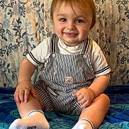 Arthur participe au concours pour gagner de l'argent avec cette photo : baby, child, smiling, striped_overalls, white_socks, blanket, cushion, pillow, patterned_fabric, indoor, seated, cute, happy, portrait, young_child, toddler, home, cozy, colorful, playful