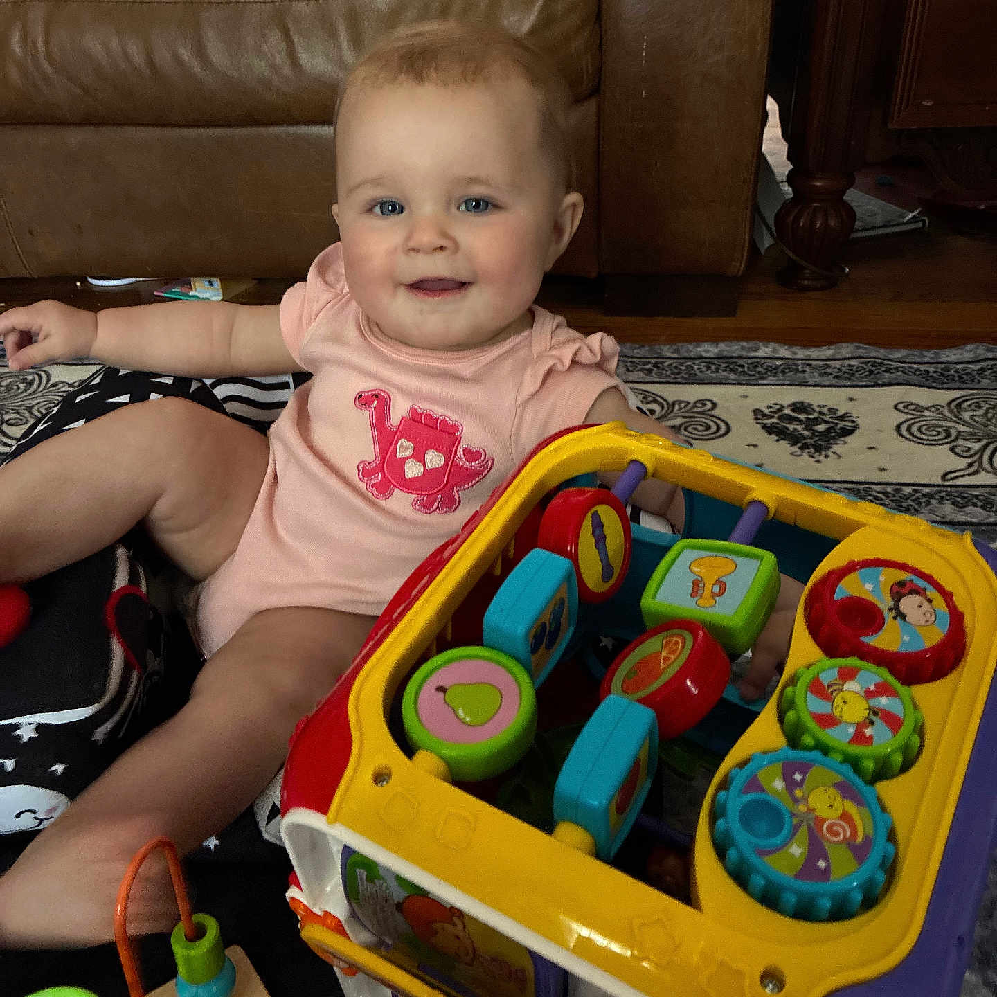 Lillian joined the competition — help win amazing prizes! baby, child, pink_onesie, dinosaur_design, toys, activity_cube, bead_maze, rug, patterned_rug, couch, leather_couch, floor, indoor, smiling, sitting, playtime, colorful, happy, person, infant