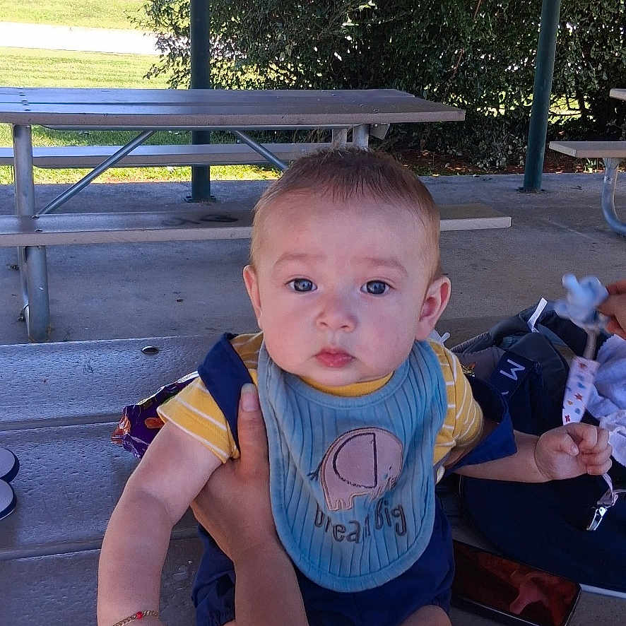 Rovin joined the competition — help win amazing prizes! baby, child, infant, outdoor, picnic, picnic_table, bench, shelter, portrait, face, blue_eyes, sitting, bib, clothing, toy, adult, legs, trees, house, daylight