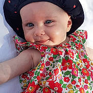 Prune a rejoint le concours — aidez-le/la à gagner de superbes lots ! baby, infant, smiling, blue_eyes, hat, black_hat, apple_pattern, floral_dress, colorful_clothing, cute, lying_down, white_blanket, cheeky_smile, child, cute_face, skin, outdoor_light, portrait, baby_fashion, happy